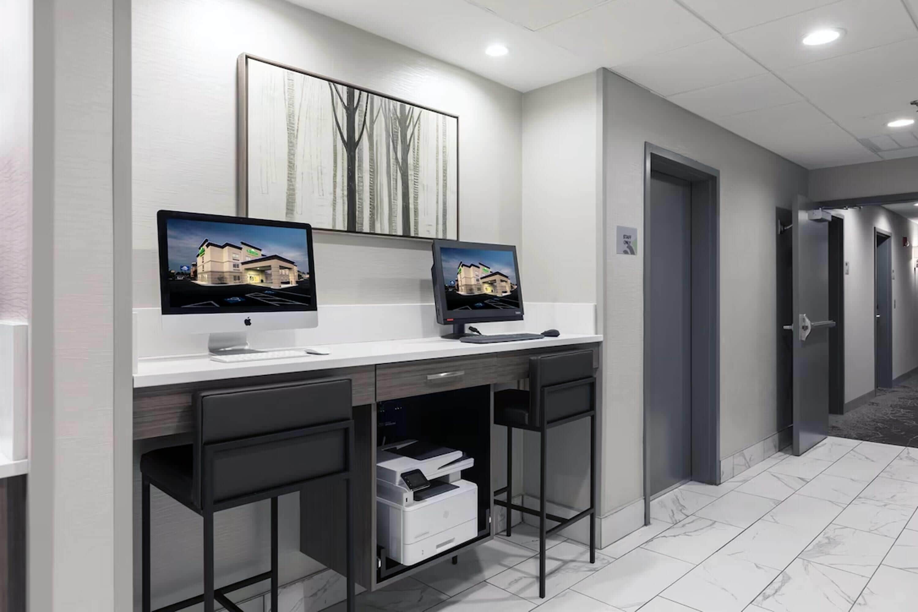 A modern office space with two desks, each with a computer monitor, a printer, and a chair. There is a large framed artwork on the wall above the desks.