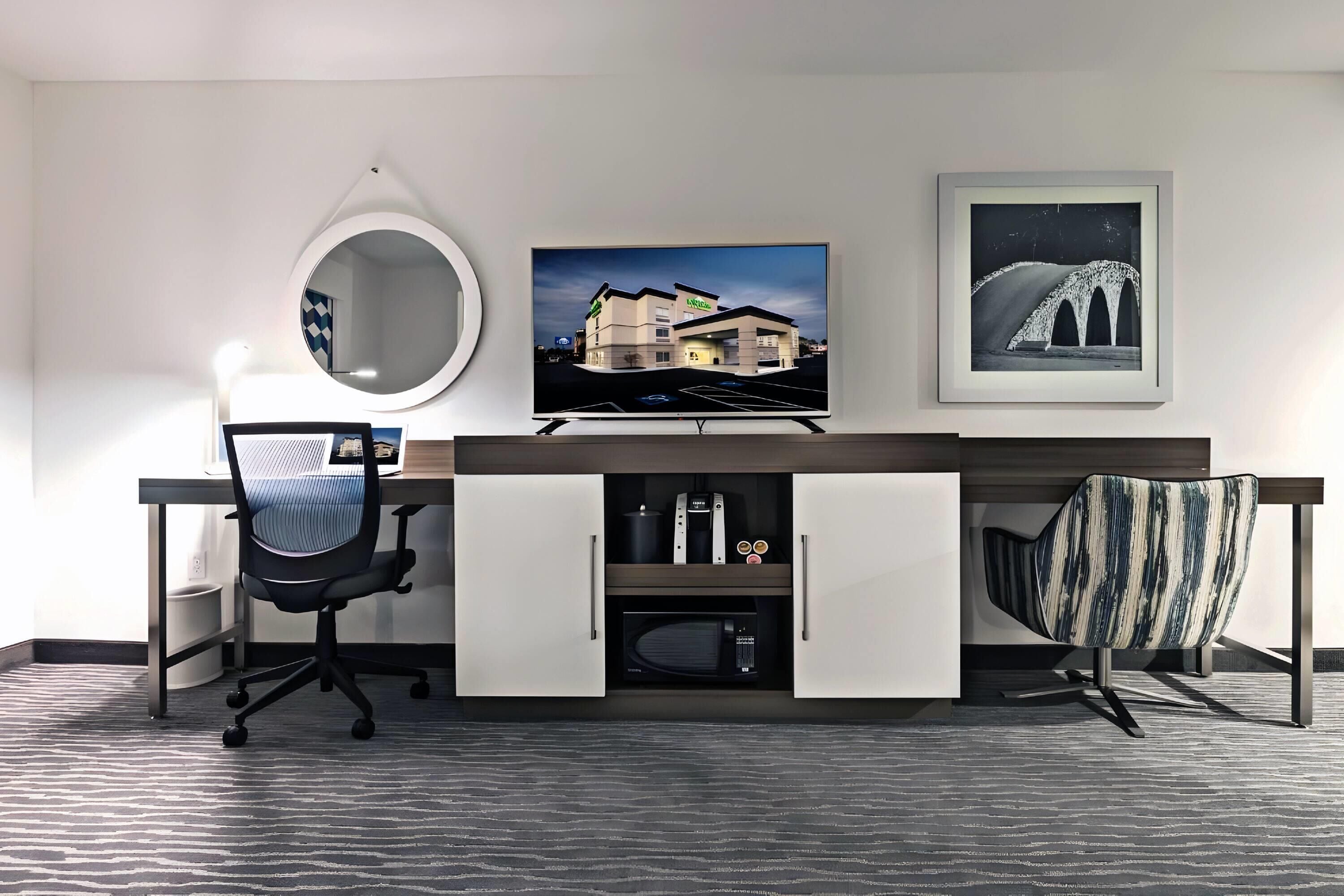 A modern hotel room with a desk, a round mirror, a flat-screen TV, and a framed picture on the wall.