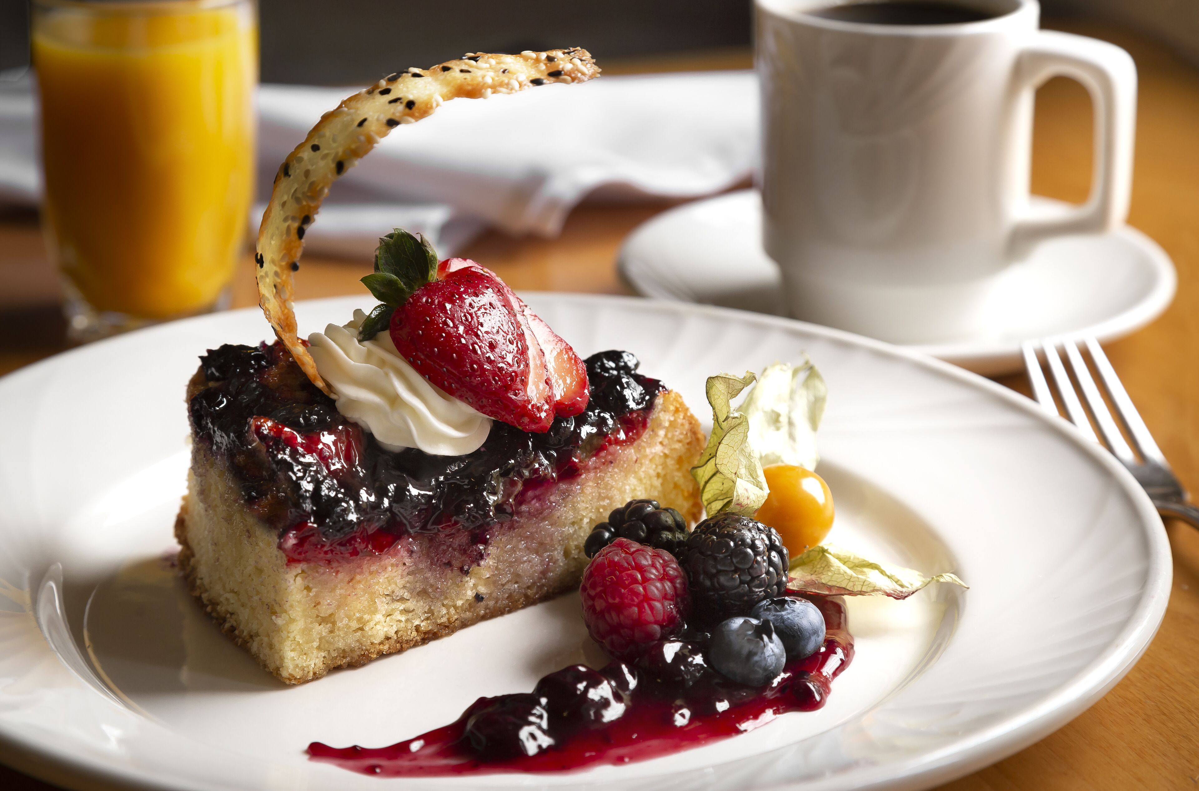 A dessert plate with a slice of cake topped with berries and whipped cream, accompanied by a glass of orange juice and a cup of coffee on a table.