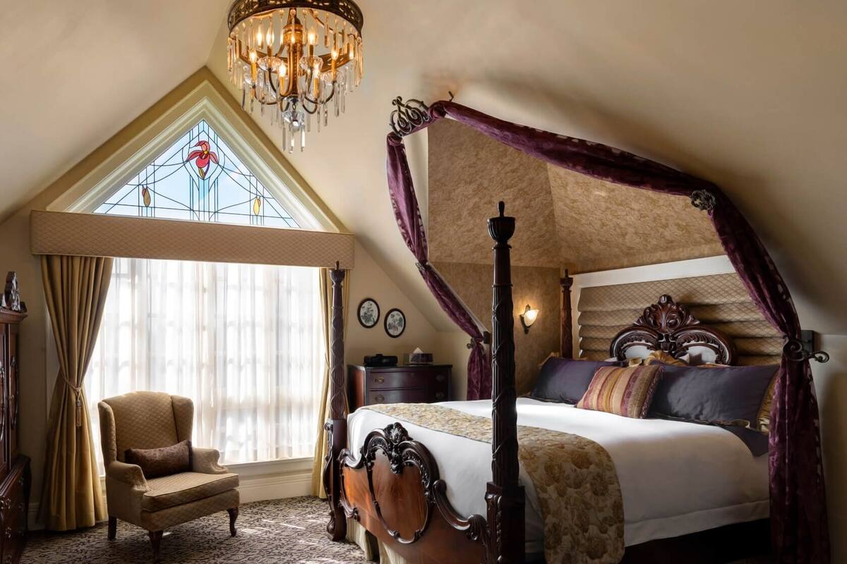 Una cama con dosel, un candelabro, una silla y un ventanal con vitrales.