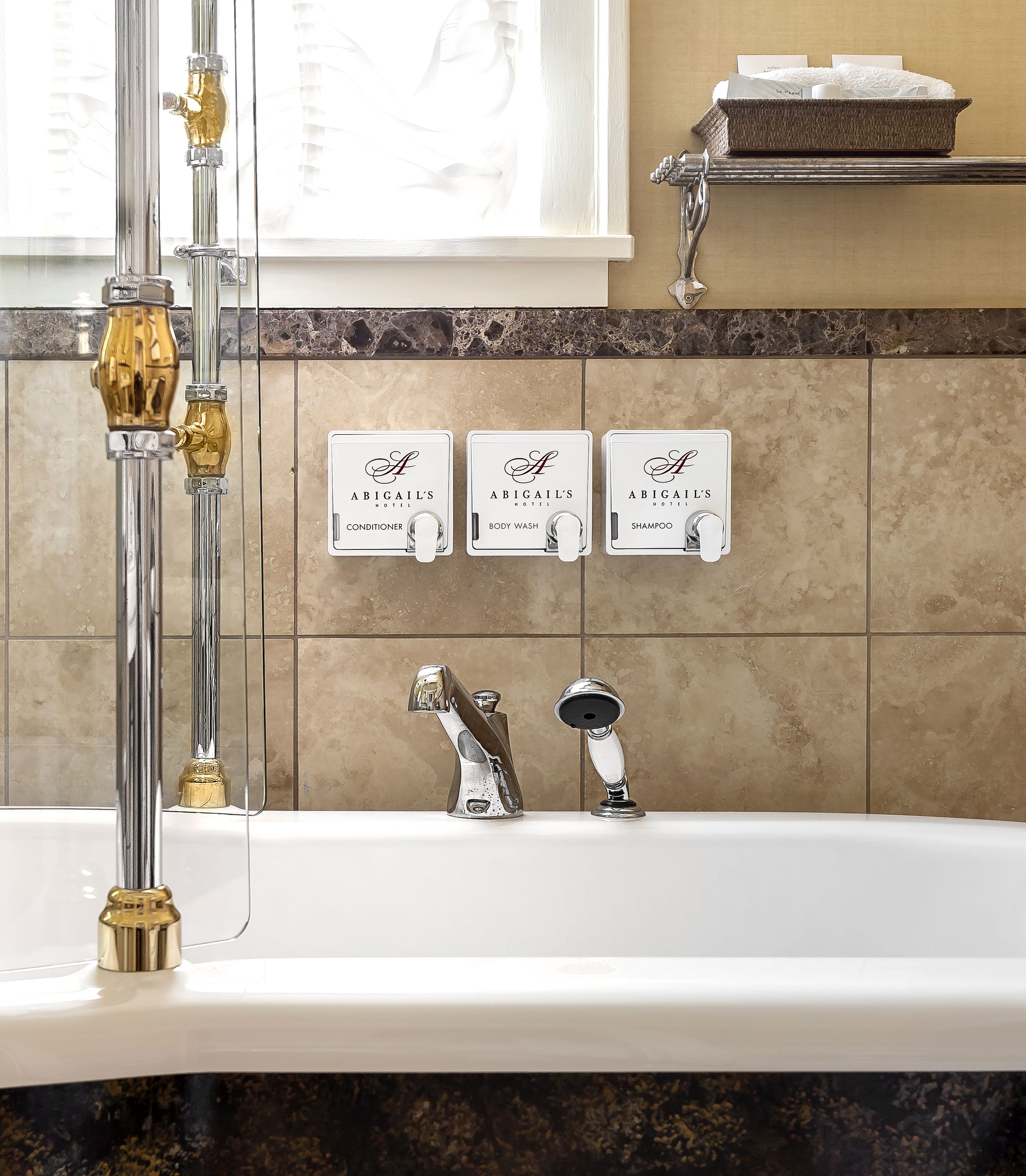 Un baño con una bañera, dos llaves y tres dispensadores montados en la pared etiquetados como “ABIGAIL'S CONDITIONER”, “ABIGAIL'S BODY WASH” y “ABIGAIL'S SHAMPOO”.