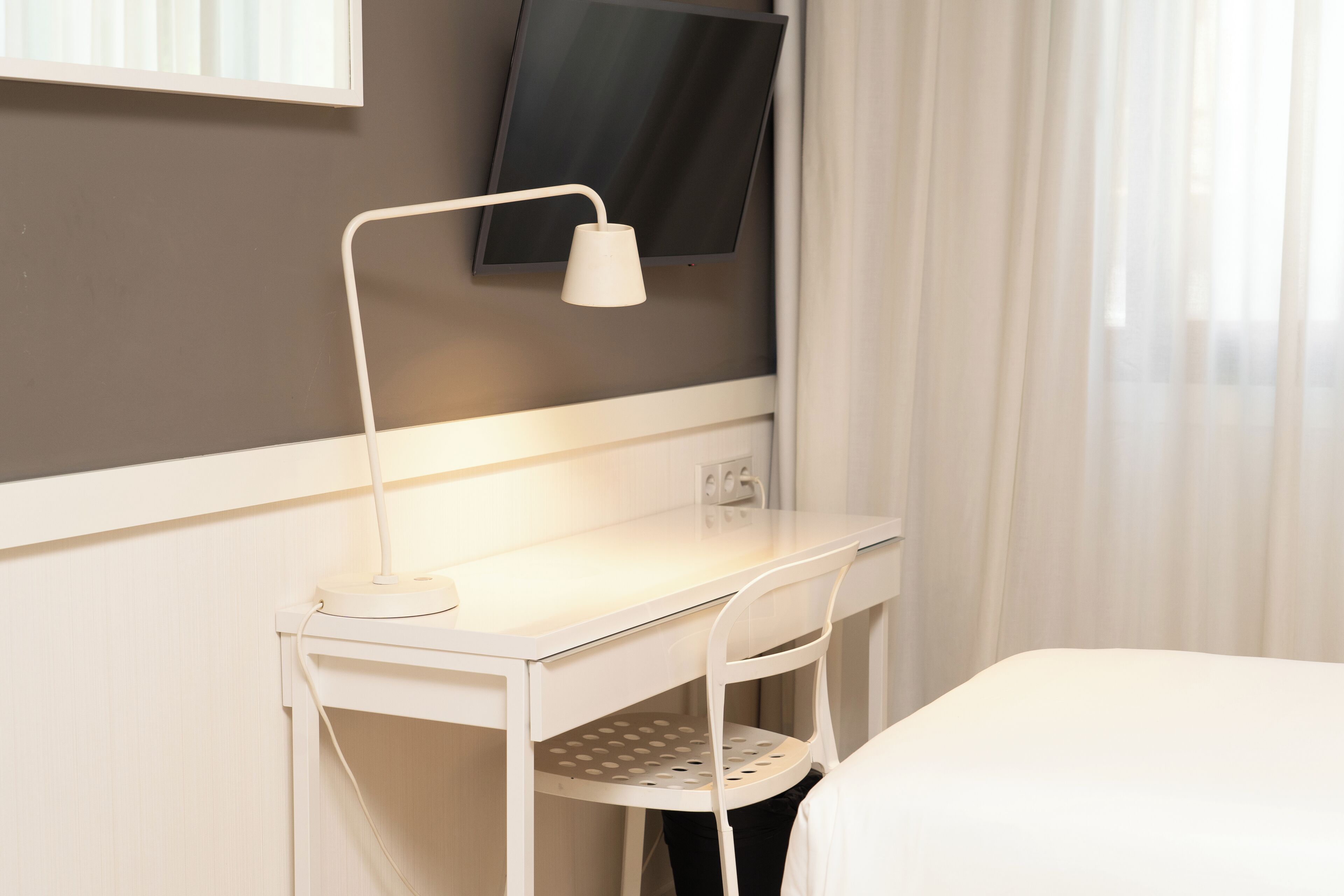 A white desk with a lamp, a TV mounted on the wall, and a bed with a white bedspread.