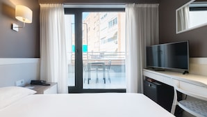 Ett hotellrum med en säng, ett skrivbord med en tv, ett litet pentry och en balkong med ett bord och stolar.