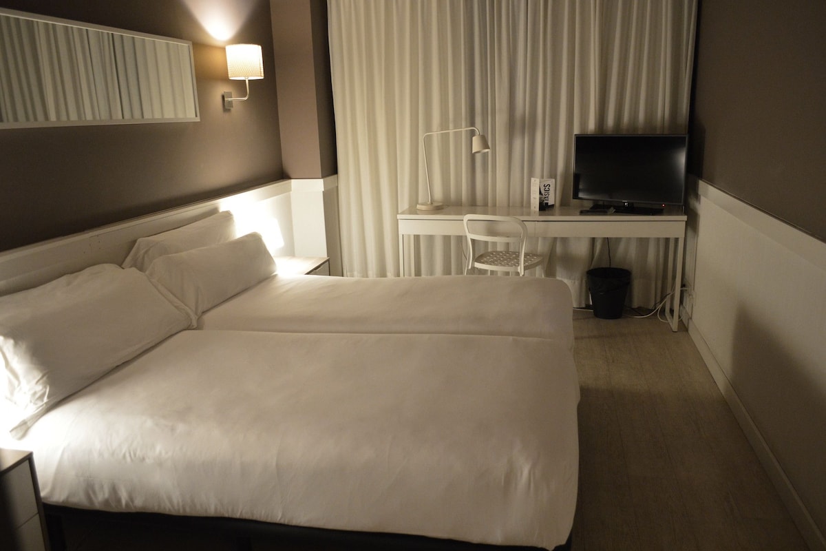 Une chambre d’hôtel avec un grand lit, un bureau équipé d’un ordinateur et d’une télévision, une chaise et une lampe fixée au mur.