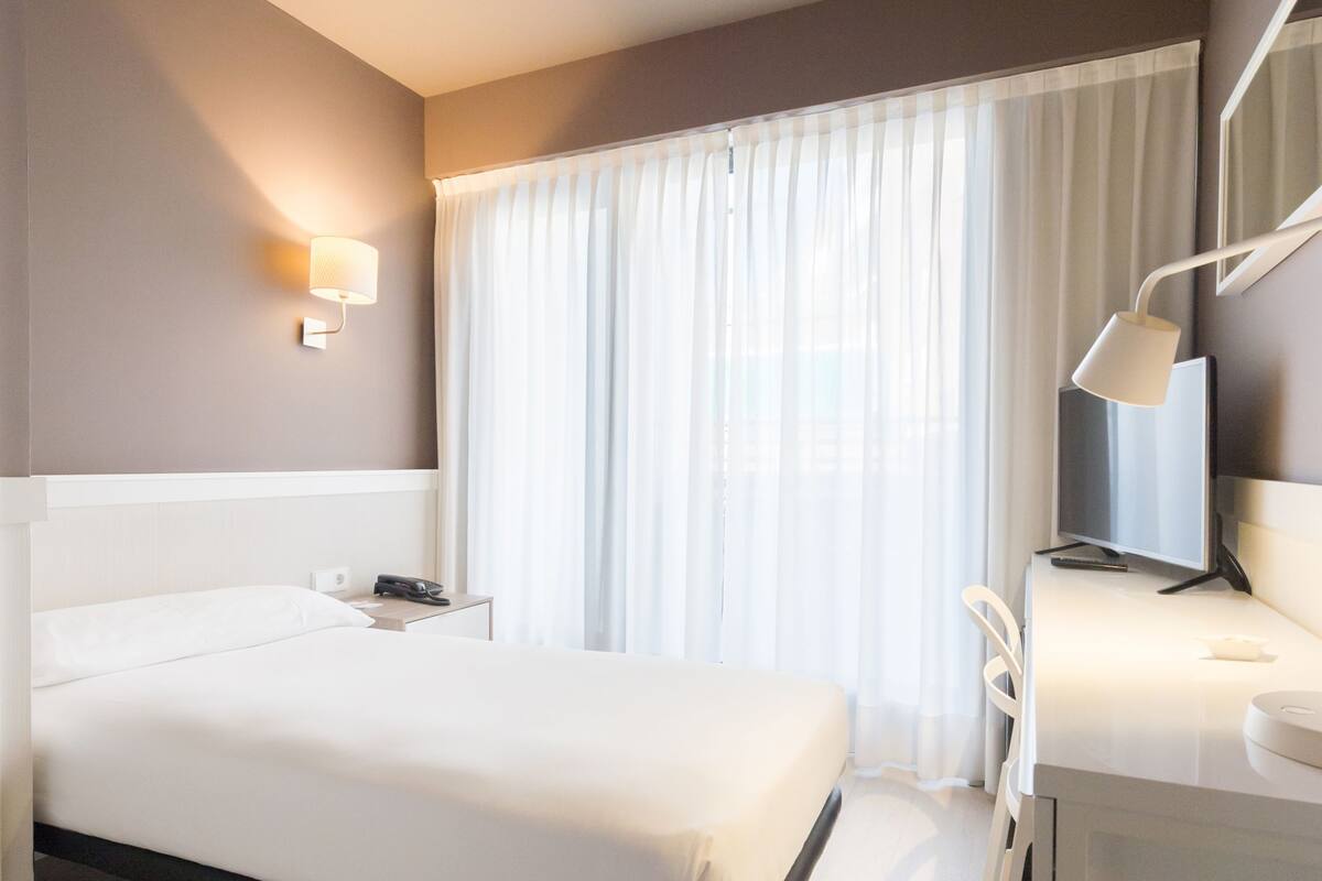 A modern hotel room with a bed, bedside table, lamp, and a desk with a chair.