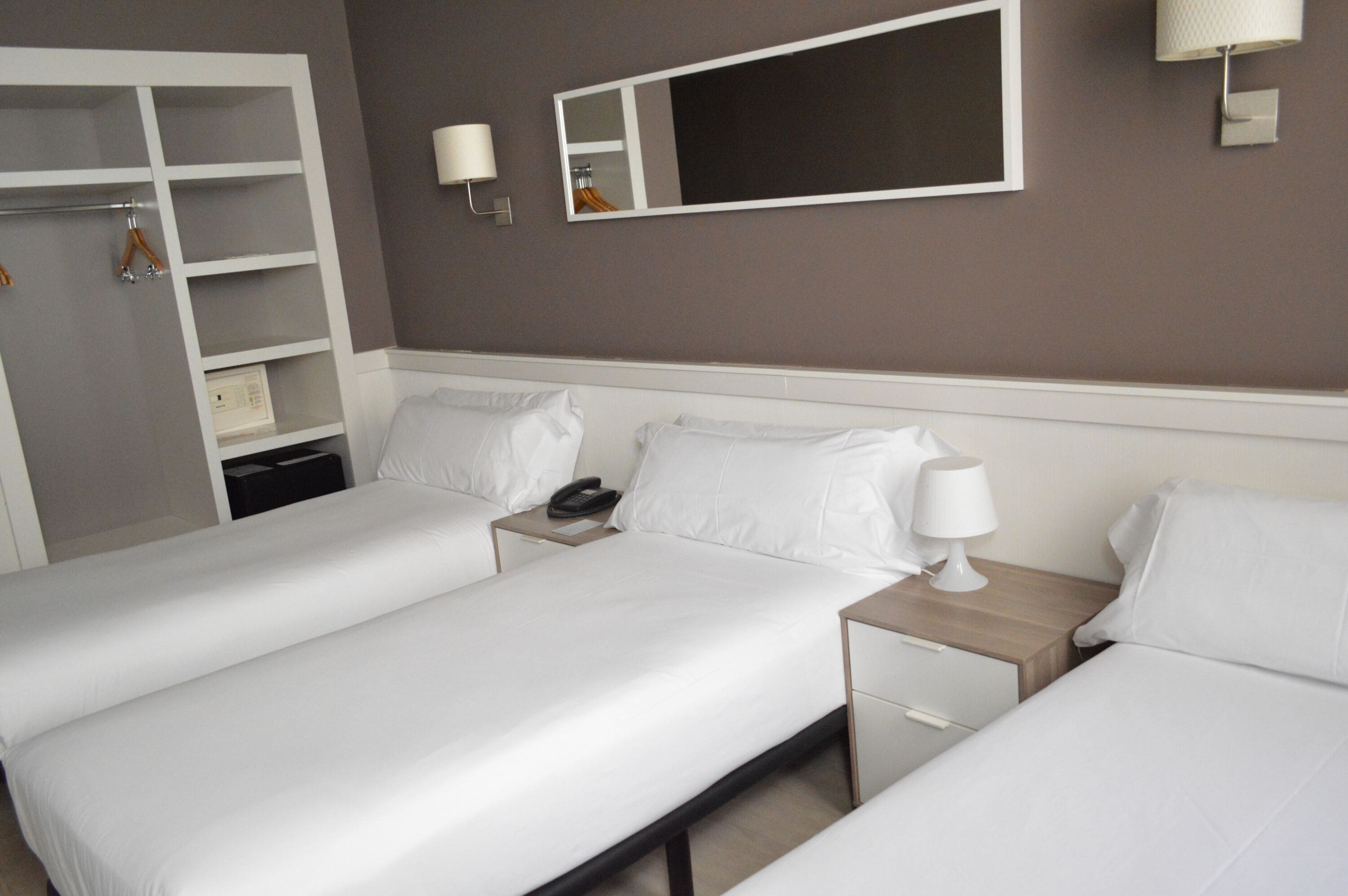 A hotel room with two single beds, a wardrobe, a bedside table with a lamp, a phone, and a mirror on the wall.