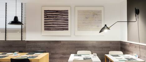 A modern dining area with tables set for meals, overhead lighting, and artwork on the walls.