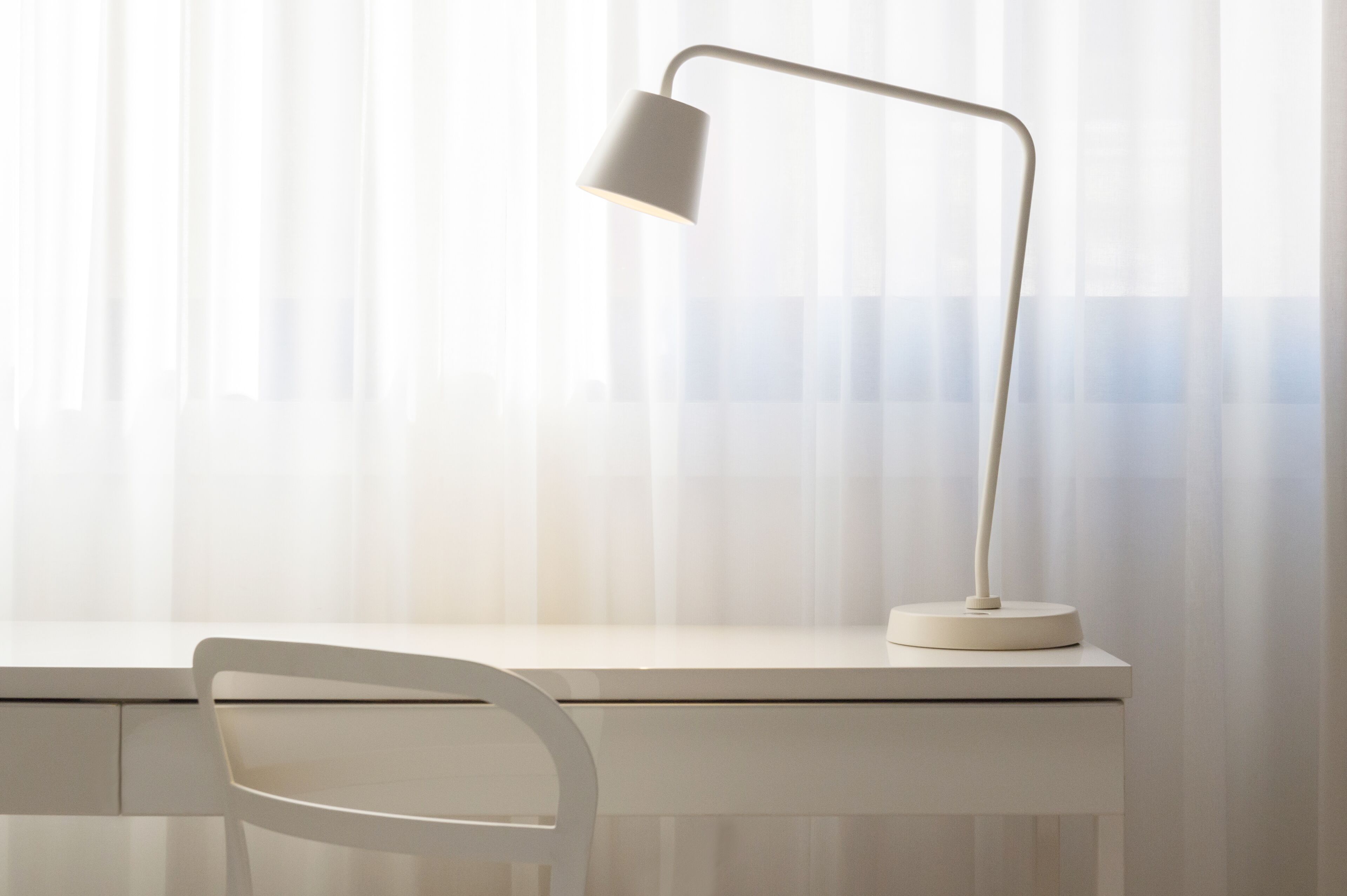 Un bureau blanc, agrémenté d’une lampe de bureau moderne et d’une chaise blanche, est placé devant une fenêtre ornée de rideaux légers.