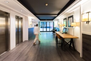 Um hall moderno de hotel com um corredor longo, elevadores, uma mesa de recepção e vista para o exterior.