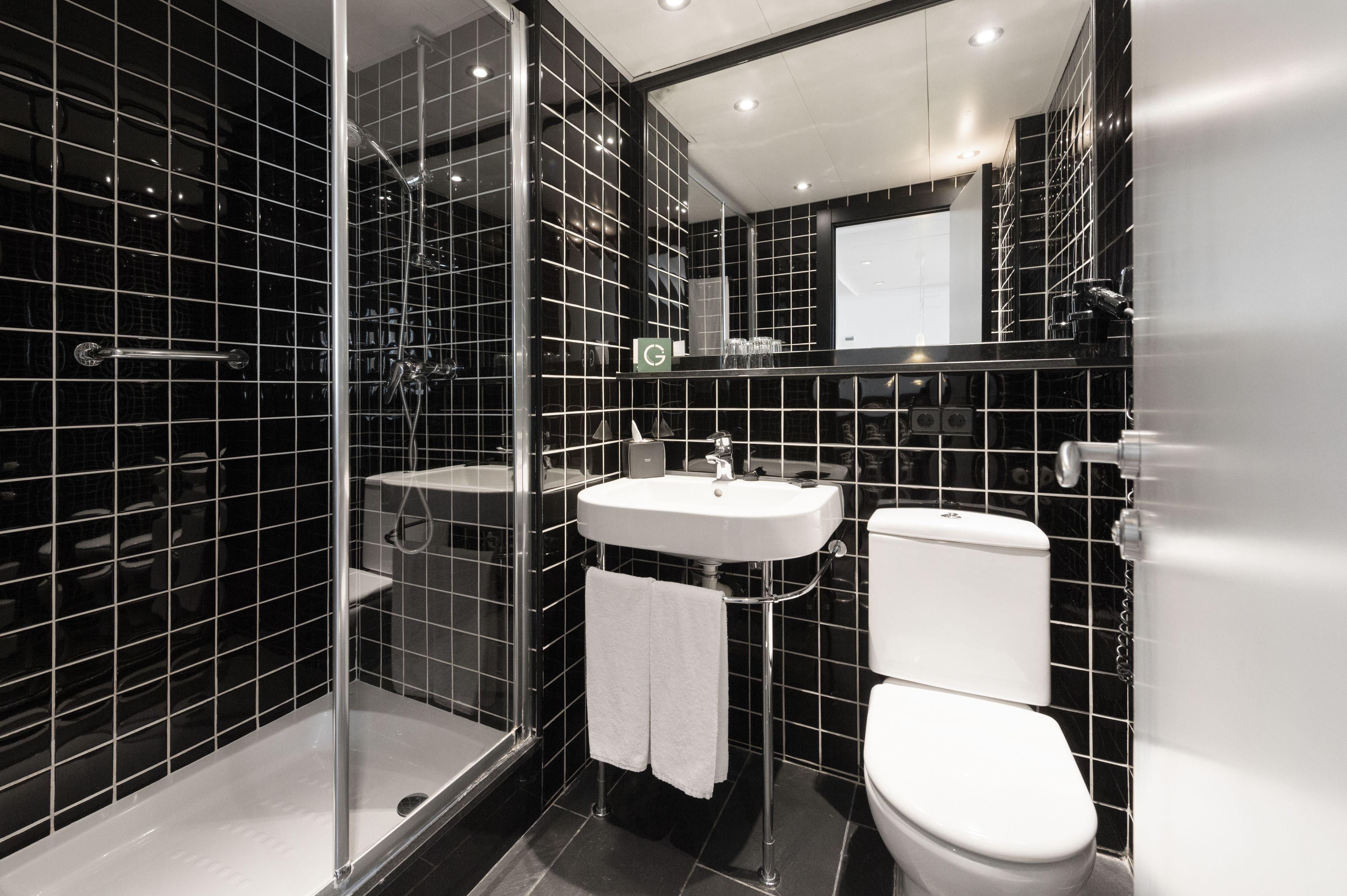 Un baño moderno con paredes de azulejos negros, cabina de ducha de vidrio, lavamanos blanco y un inodoro.