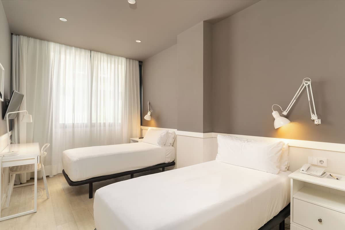 A hotel room with two beds, a desk, a chair, and a bedside table with a lamp.