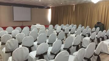 Un salón de conferencias con filas de sillas cubiertas de blanco y atadas con cintas marrones, una pantalla de proyector y un atril.
