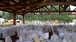 Un salón de banquetes al aire libre con varias mesas dispuestas para un evento formal, rodeado por una estructura de pérgola de madera.