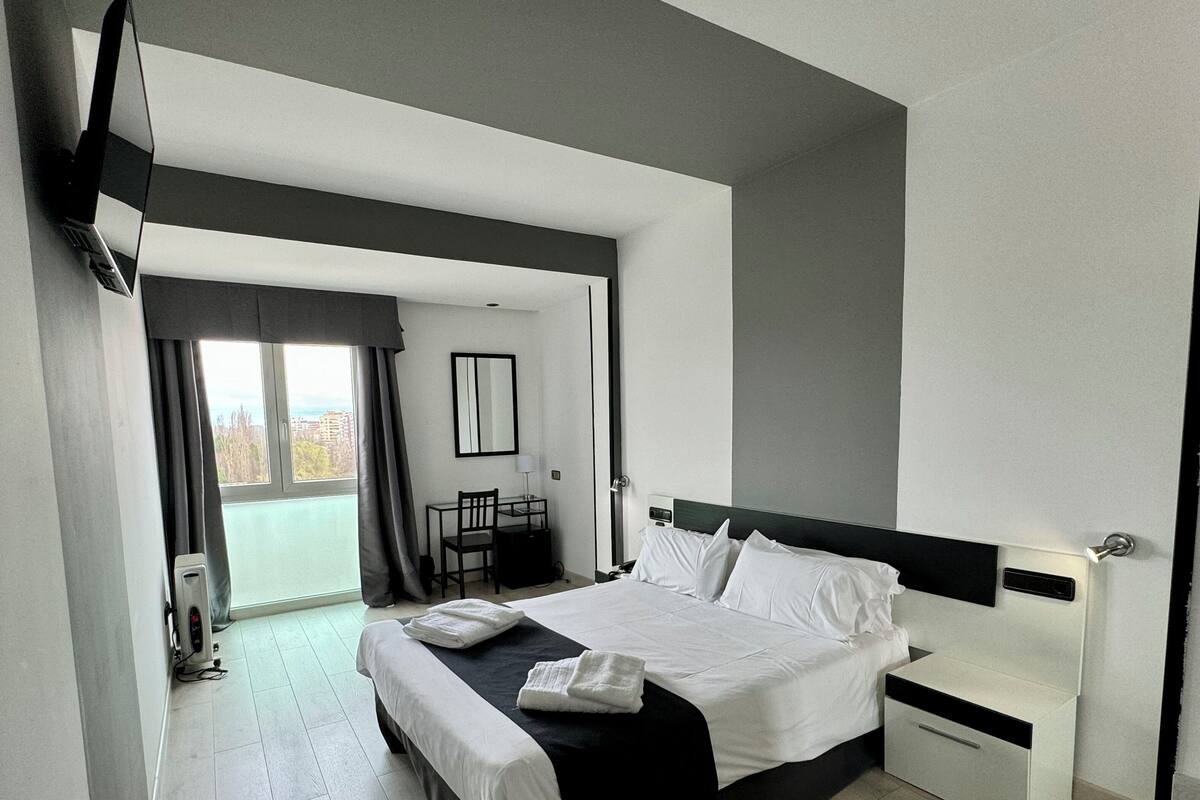 A modern hotel room with a large bed, a bedside table, a desk with a chair, and a view of the city through the window.