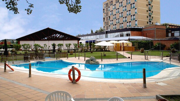 Un hotel con piscina, mobiliario exterior y un edificio grande al fondo.