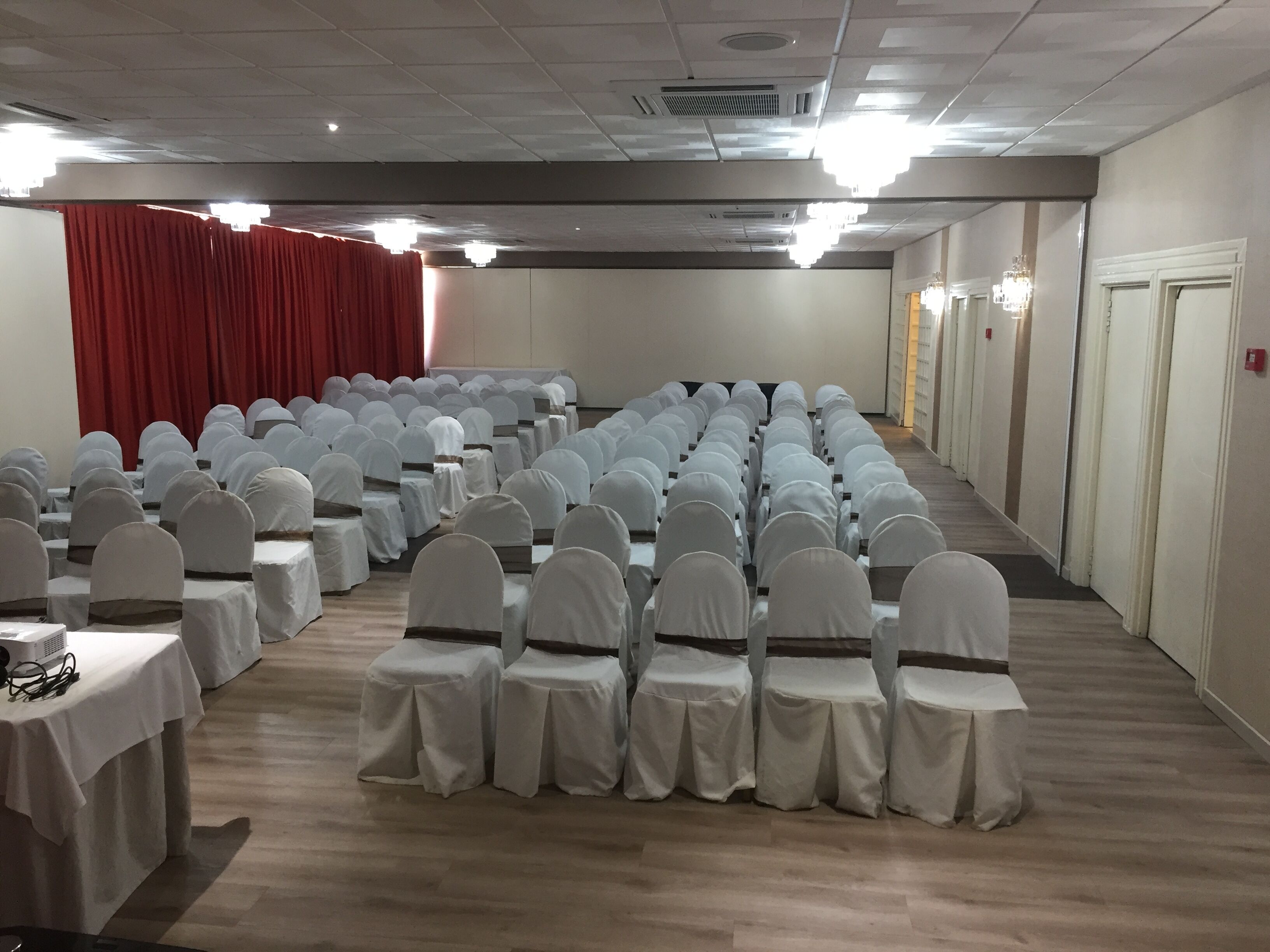 Un salón de conferencias con filas de sillas y mesas cubiertas de blanco, un telón de fondo rojo y piso de madera.
