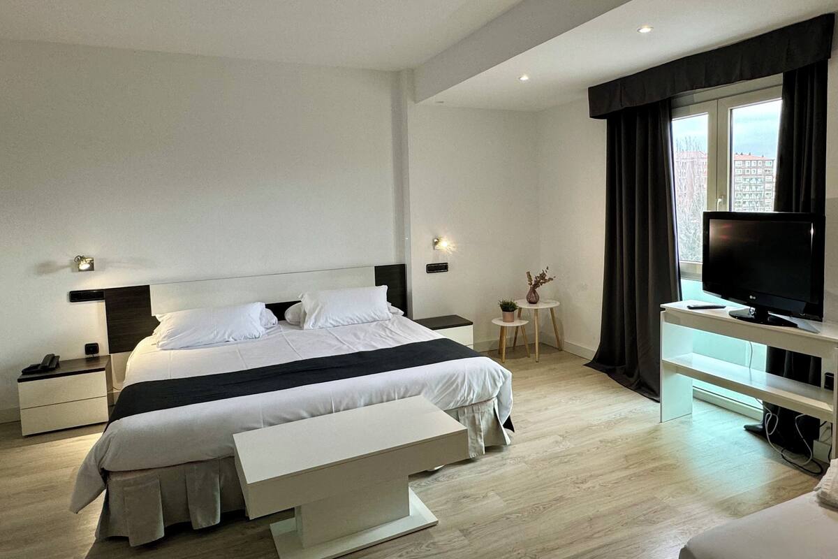 A modern hotel room with a large bed, bedside tables, a TV, and a small side table with a plant.