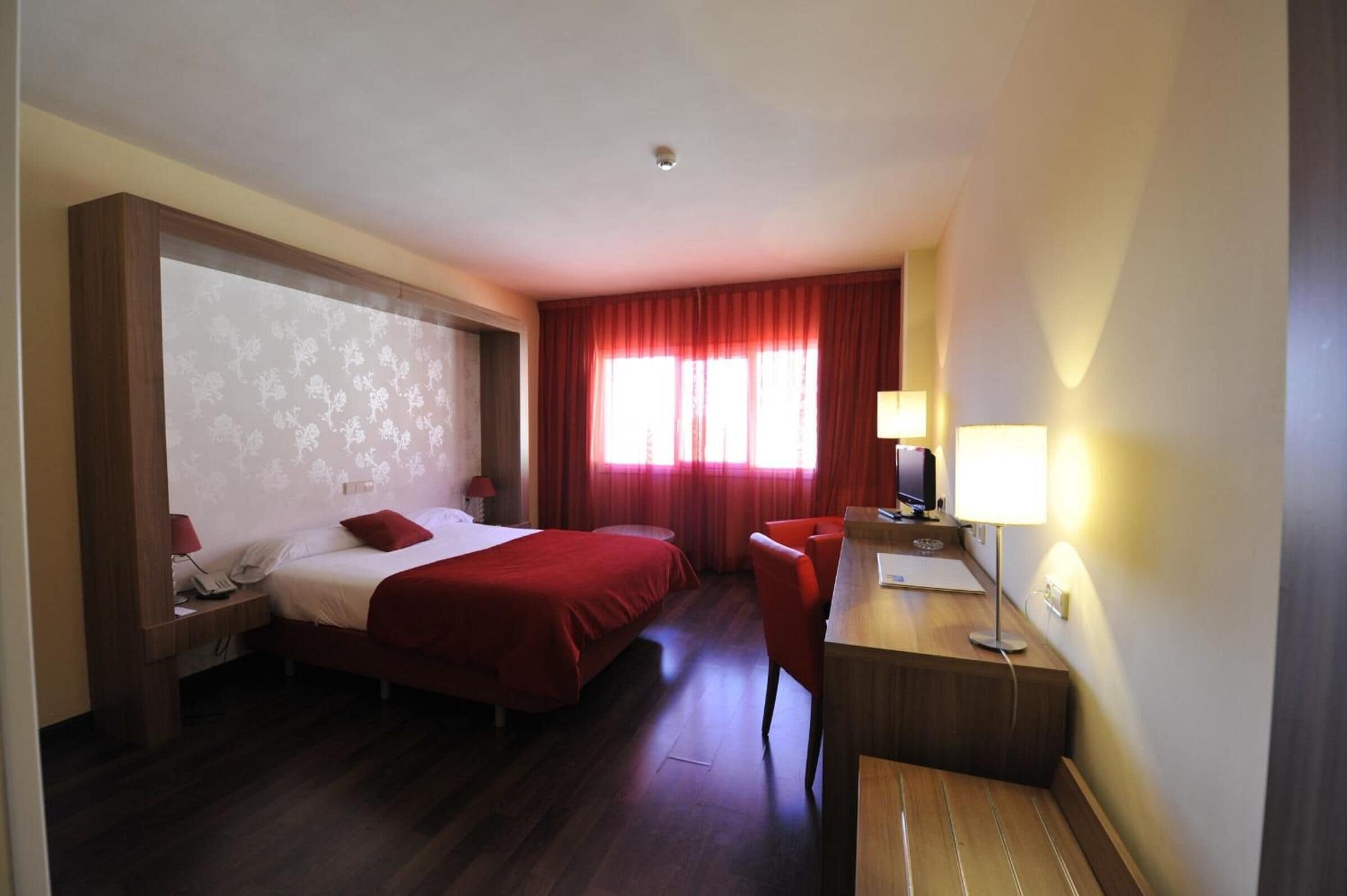 A hotel room with two beds, a desk, a chair, a desk lamp, and a window with red curtains.