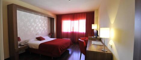 Habitación de hotel con dos camas, un escritorio, una silla, una lámpara de escritorio y una ventana con cortinas rojas.