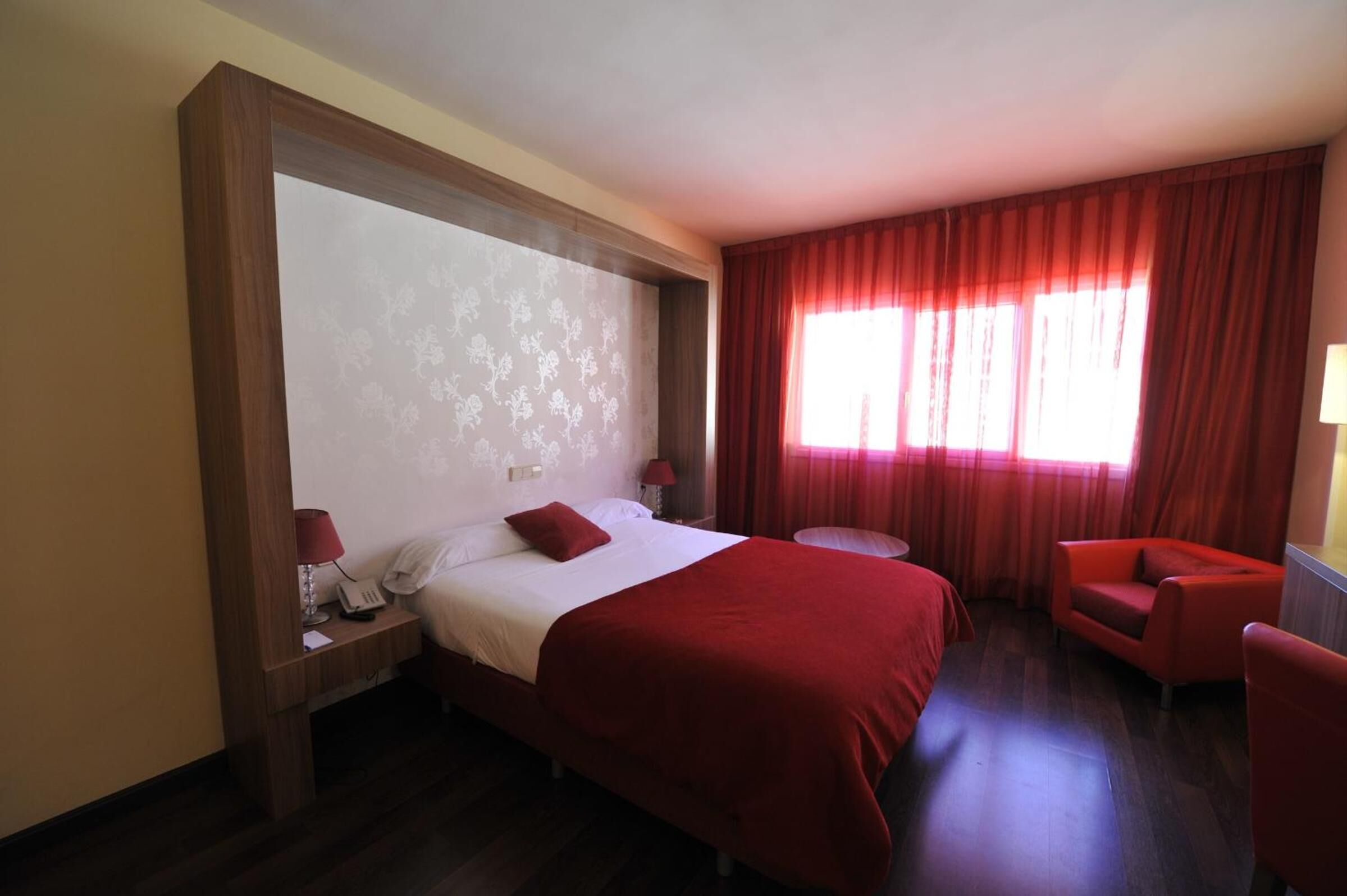 Habitación de hotel con una cama grande, una mesita redonda pequeña, un sillón rojo y una ventana con cortinas rojas.