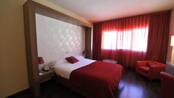 Habitación de hotel con una cama grande, una mesita redonda pequeña, un sillón rojo y una ventana con cortinas rojas.