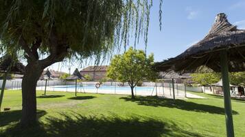 A pool area with a thatched-roof structure, a tree, and a clear blue sky.
