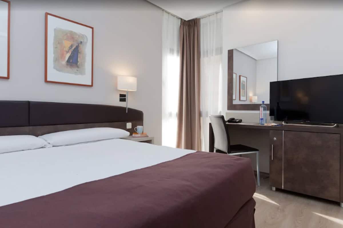 A hotel room with a large bed, a desk with a television, a chair, a desk lamp, and framed artwork on the wall.