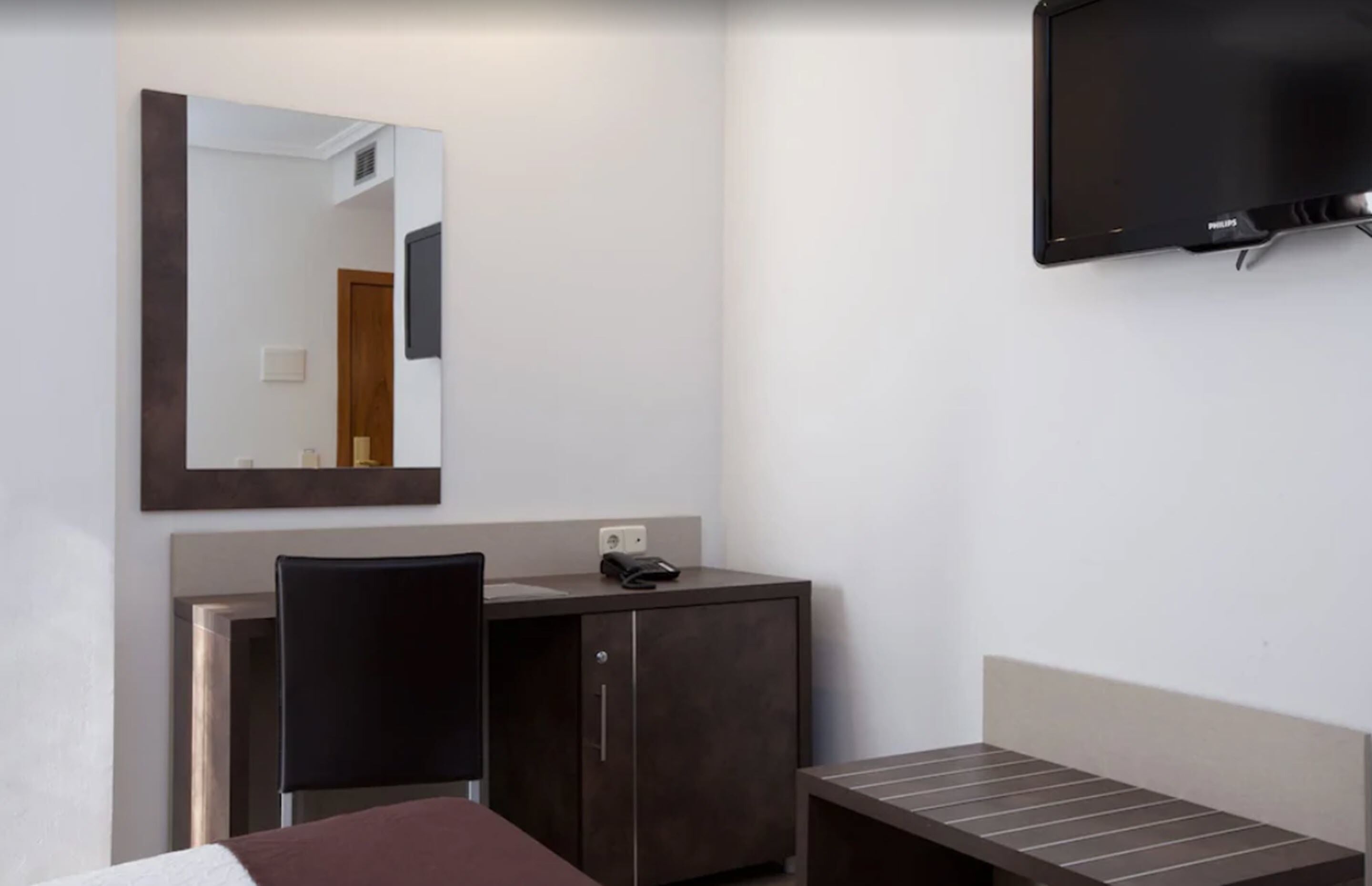 Una habitación de hotel con una cama, un escritorio con silla, un mueble pequeño, un televisor montado en la pared y un espejo sobre el escritorio.