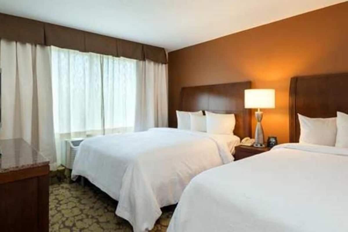 Una camera d'albergo con due letti, un comodino con lampada, un telefono e una finestra con tende.
