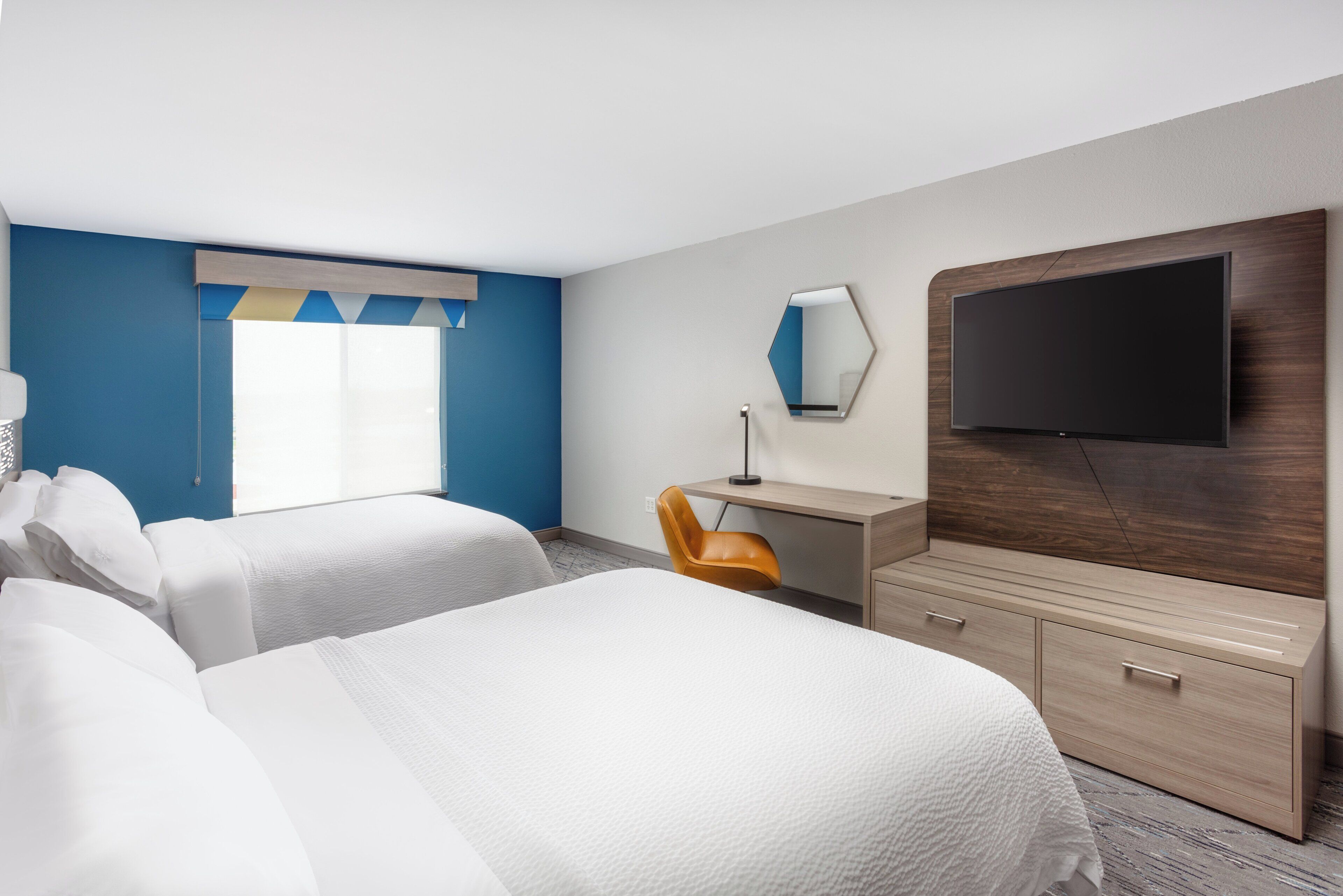 A hotel room with two beds, a desk, a chair, a flat-screen TV, and a large window with curtains.