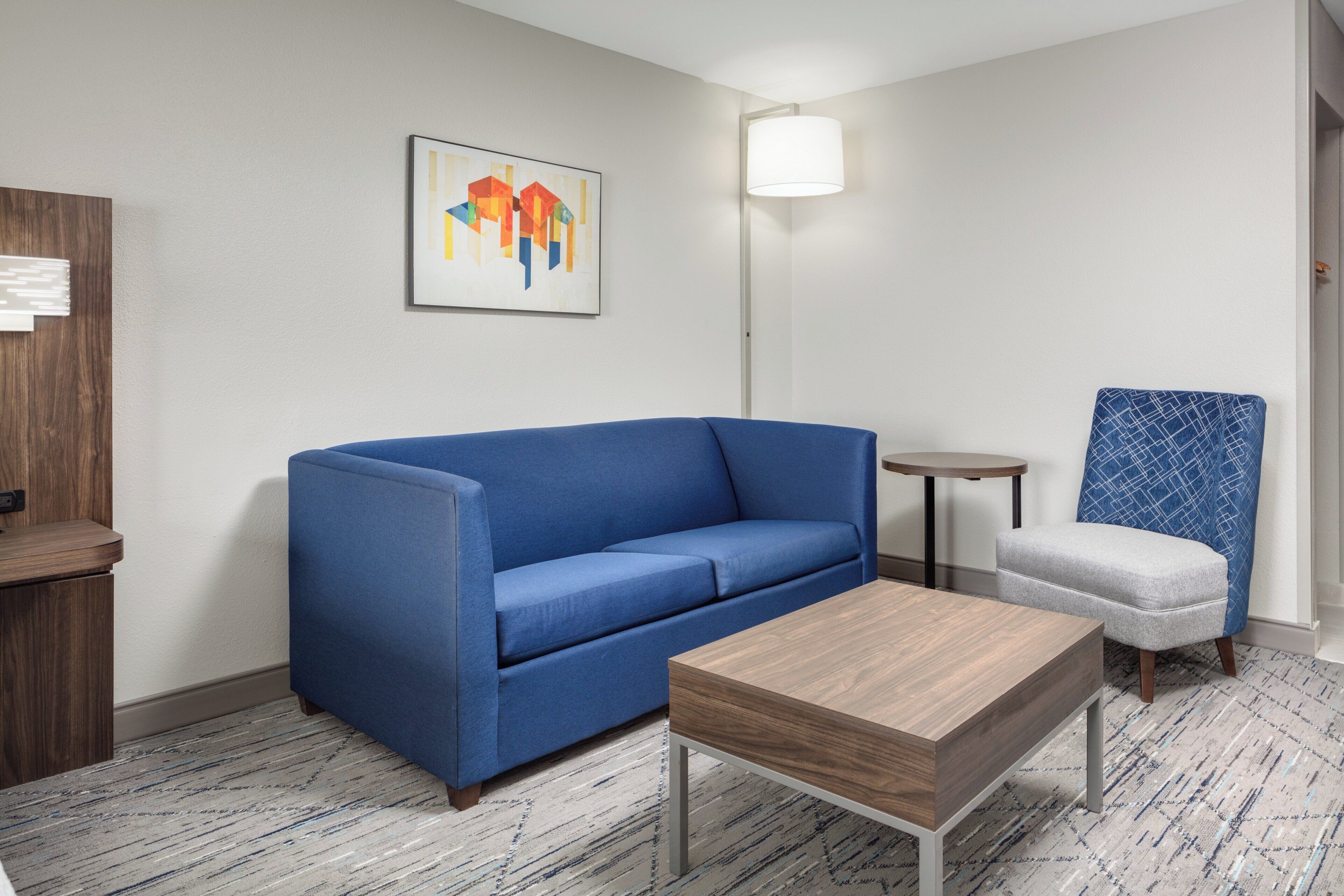 A modern hotel room with a blue sofa, a wooden coffee table, a small round table, a patterned armchair, and a painting on the wall.