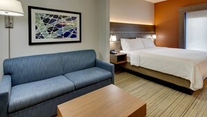 A hotel room with a blue sofa, a wooden coffee table, a bed with white linens, a nightstand with a lamp, and a framed abstract artwork on the wall.