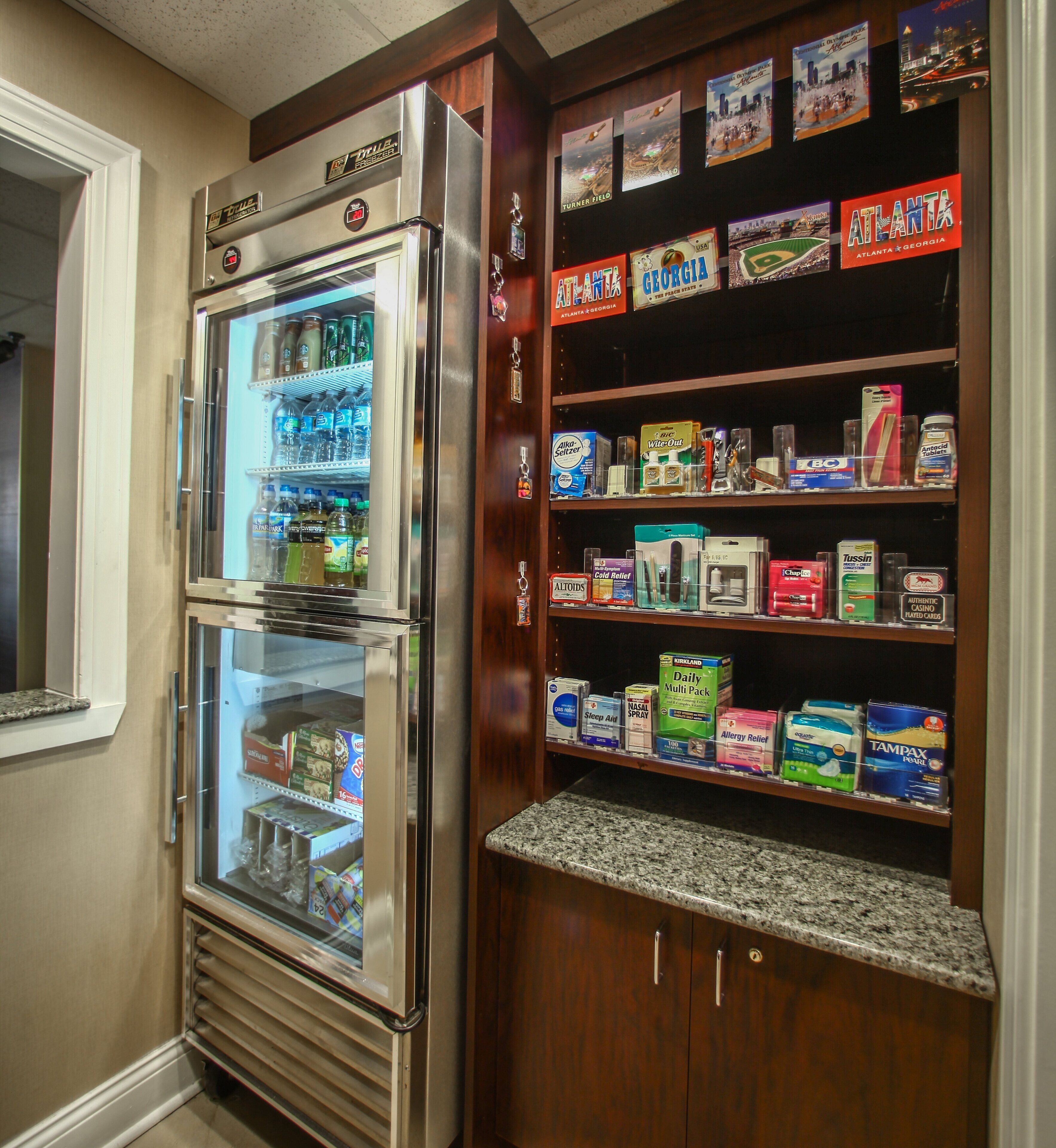 A double-door refrigerator with a freezer compartment, stocked with various beverages and food items. Adjacent to it is a wooden shelf unit displaying an assortment of products including beverages, personal care items, and household goods.