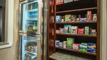 A double-door refrigerator with a freezer compartment, stocked with various beverages and food items. Adjacent to it is a wooden shelf unit displaying an assortment of products including beverages, personal care items, and household goods.