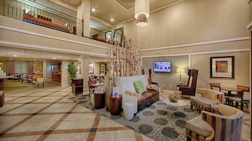 Un hall d’entrée spacieux avec un plafond haut, un espace de détente central avec un canapé et des fauteuils, un escalier et un écran de télévision fixé au mur.