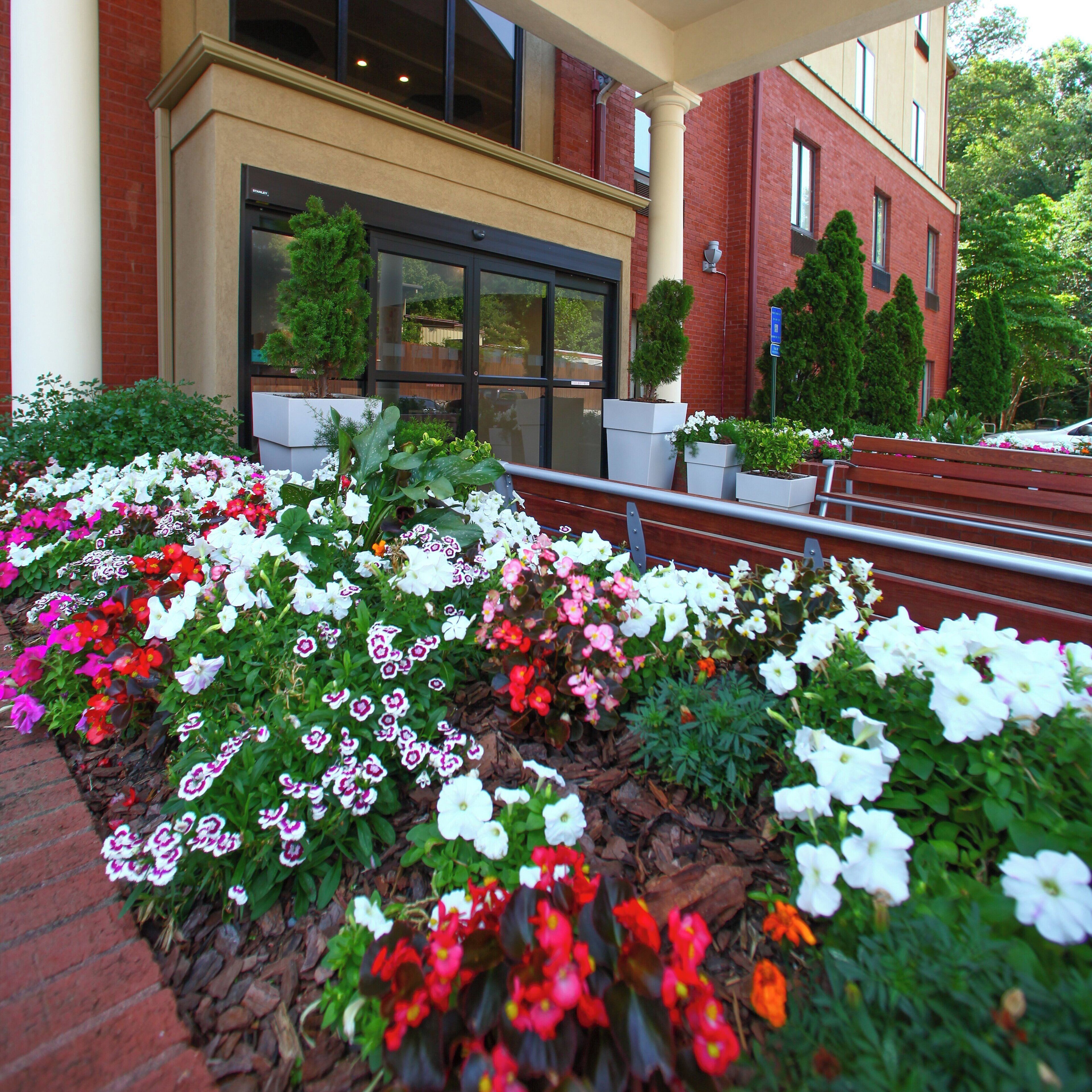 A building entrance with a brick facade, large windows, and a flower bed with various flowers.