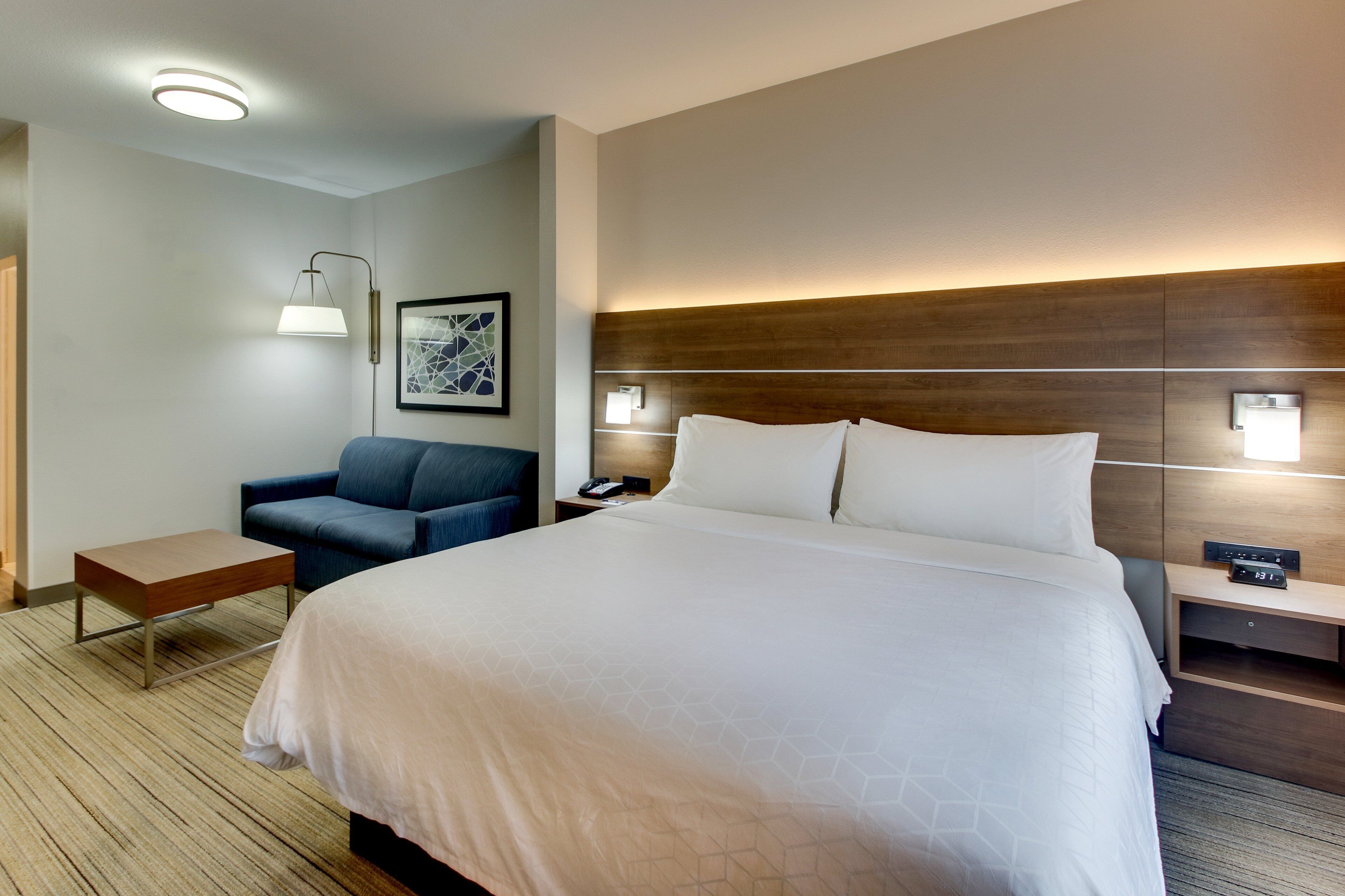 Een hotelkamer met een groot bed, een nachtkastje, een stoel, een lamp en een wandlamp.