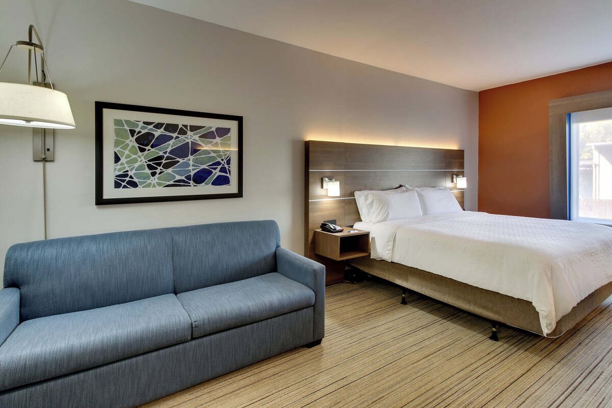 Une chambre d’hôtel comprenant un lit, un canapé, une table de chevet, une lampe fixée au mur et un tableau abstrait encadré.