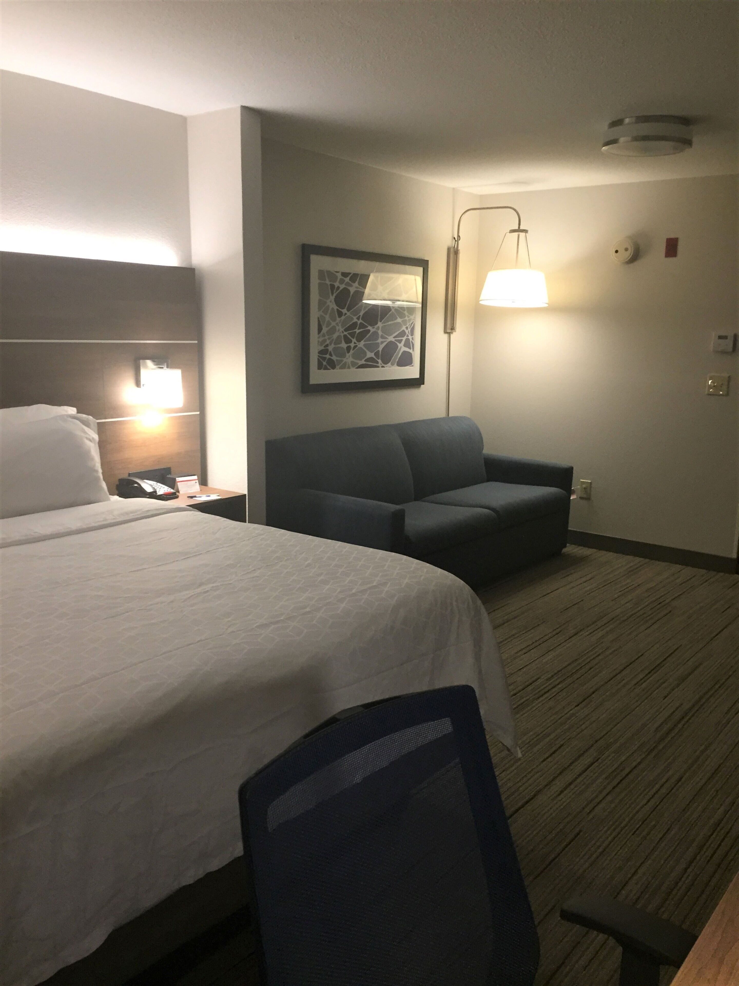 A hotel room with a large bed, a desk, a chair, a sofa, a framed artwork, and a wall-mounted lamp.