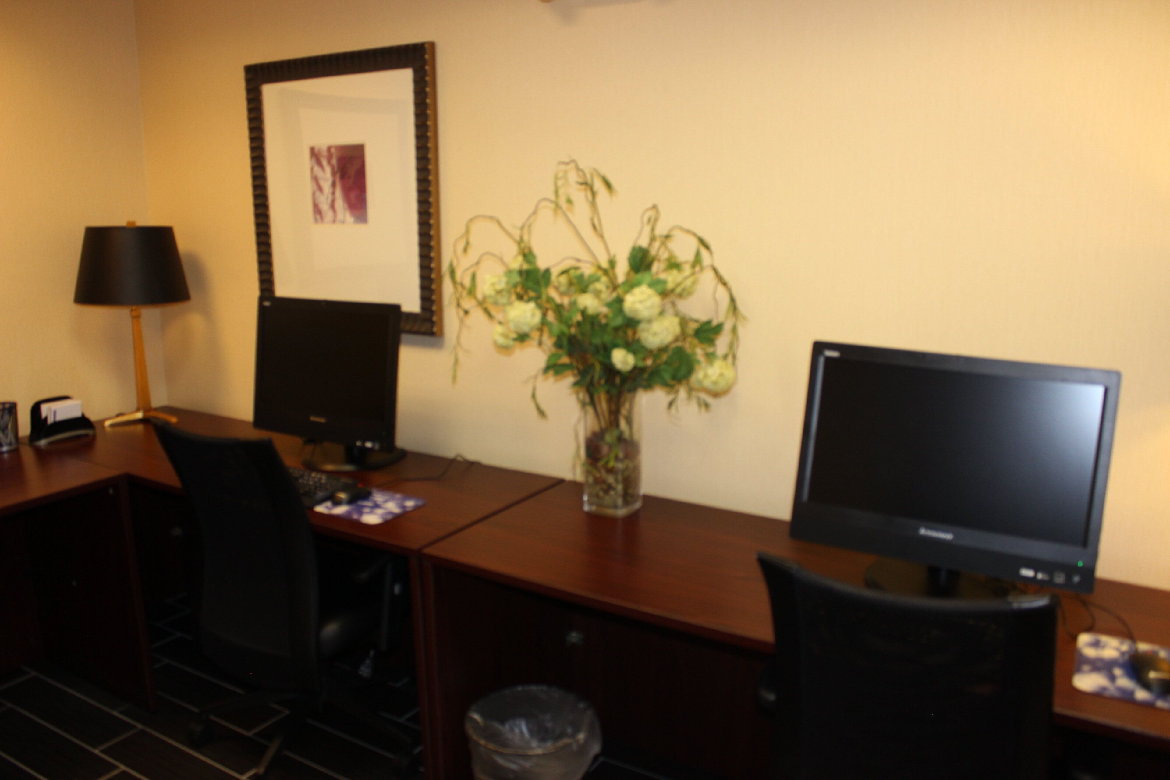 Habitación de hotel con un escritorio, dos monitores, una silla, una lámpara y un jarrón con flores.