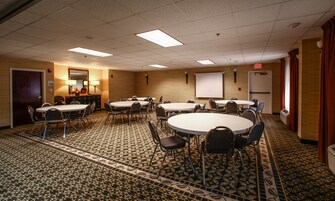 Une salle de conférence dotée de plusieurs tables et chaises, d’un écran de projection et d’un sol recouvert de moquette.