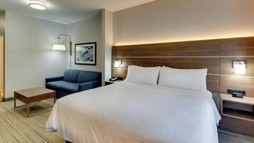 A hotel room with a large bed, a desk, a chair, a bedside table, a lamp, a framed picture, and a wall-mounted phone.