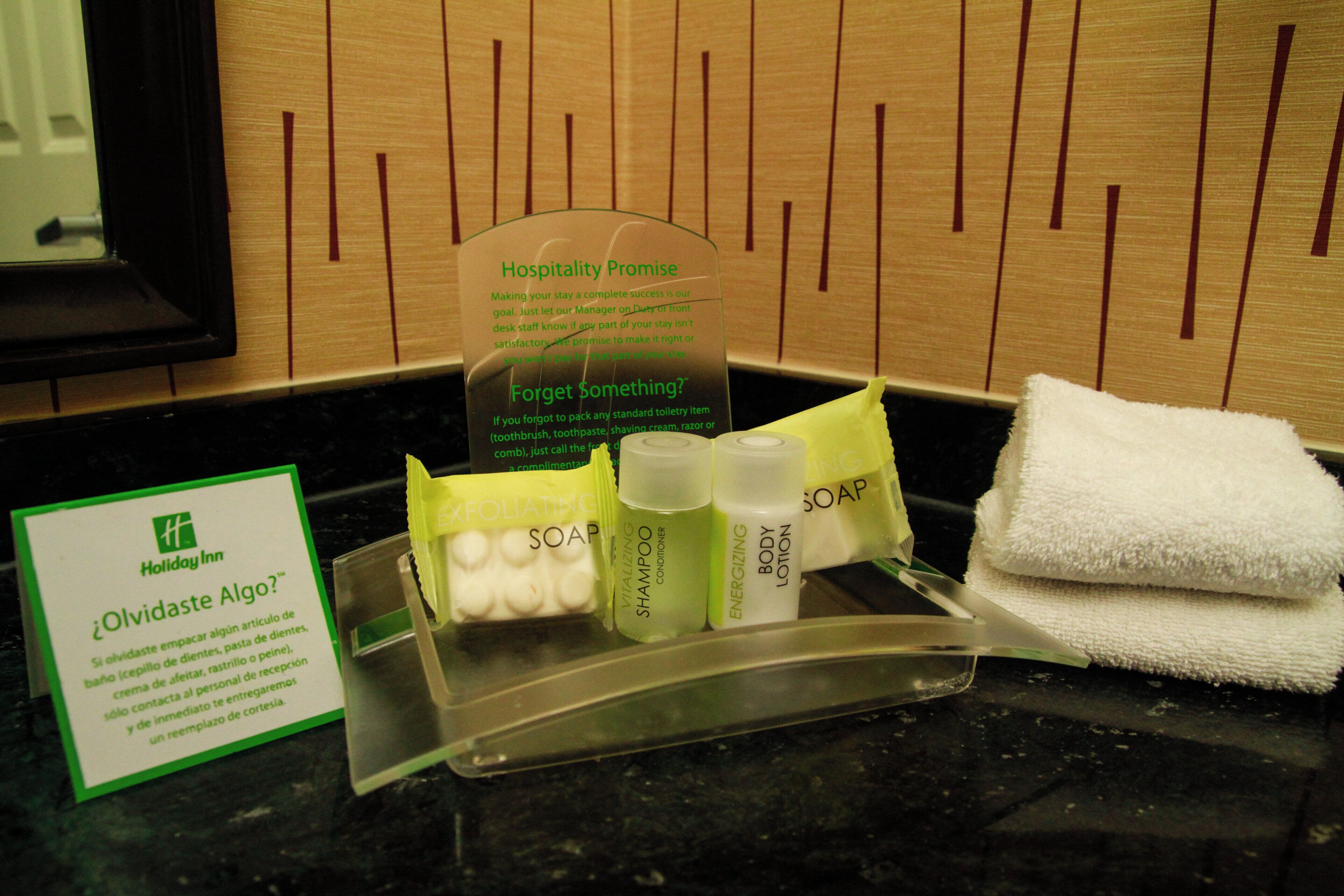 A hotel bathroom counter with toiletries, a towel, and a sign offering hospitality promise.