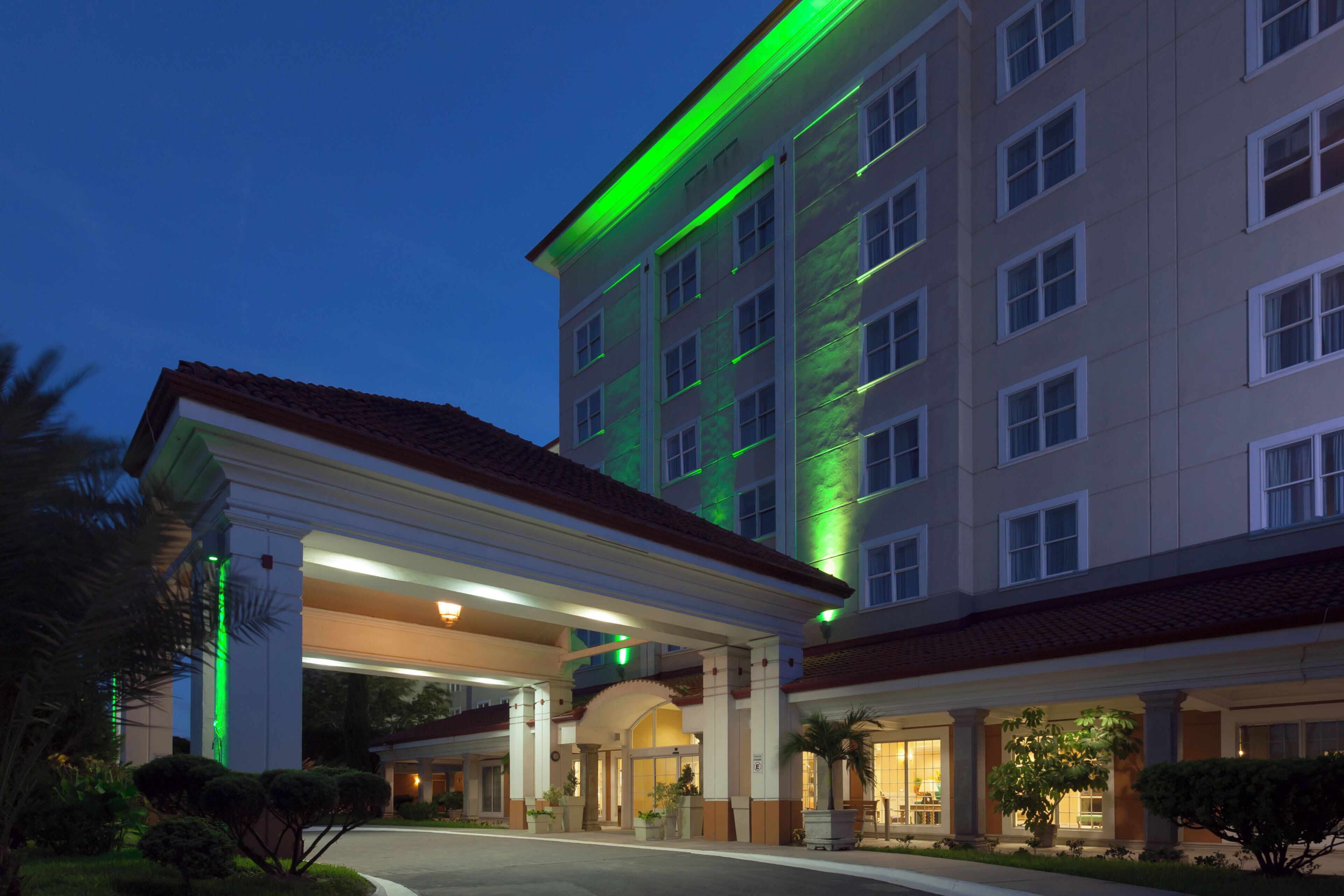 Un hotel de varios pisos con fachada iluminada en verde, un acceso cubierto y un camino de entrada ajardinado.