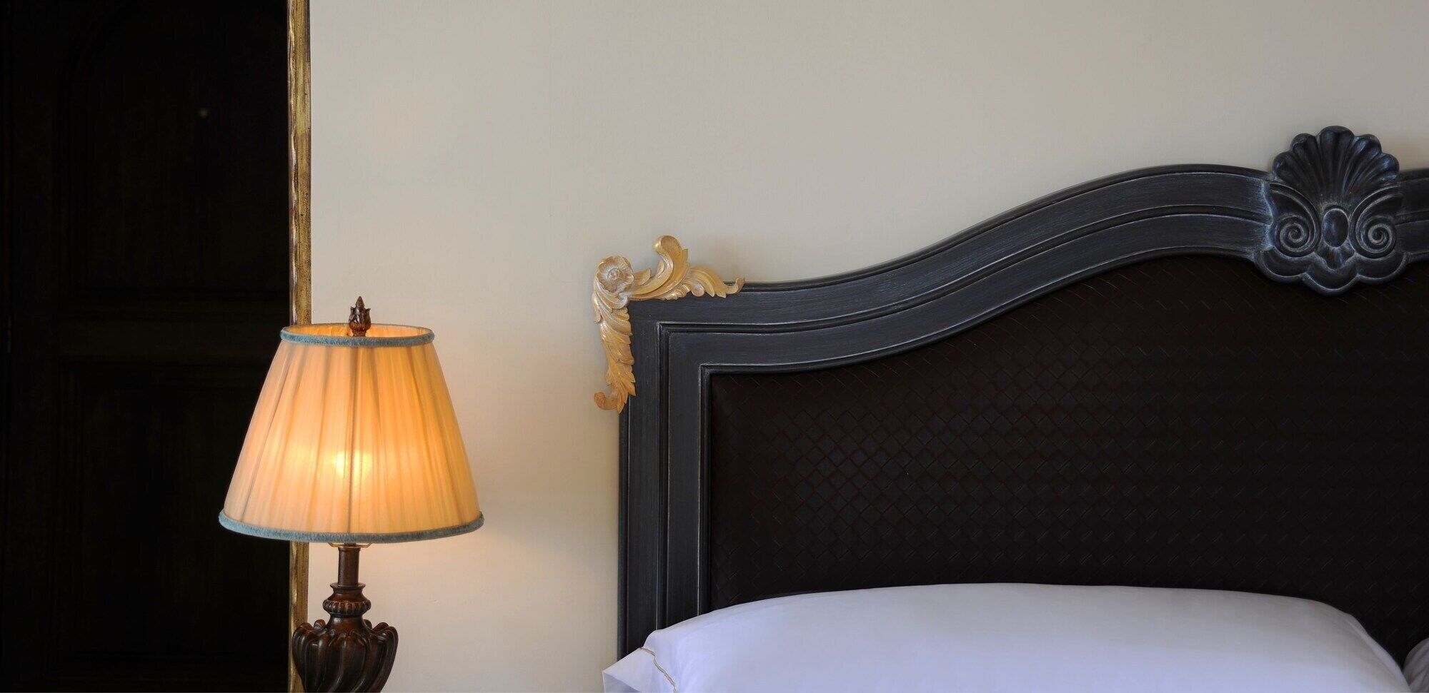 Una cama con cabecera oscura y almohada blanca, una lámpara con pantalla beige y una pared con detalles dorados decorativos.