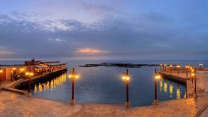 A waterfront promenade with a stone pathway, a wooden deck, and a row of lights along the water's edge. - Byblos Sur Mer (Byblos)