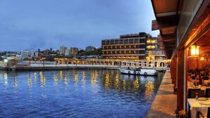 A waterfront restaurant with outdoor seating overlooking the water, a boat docked nearby, and a building with multiple balconies in the background. - Byblos Sur Mer (Byblos)