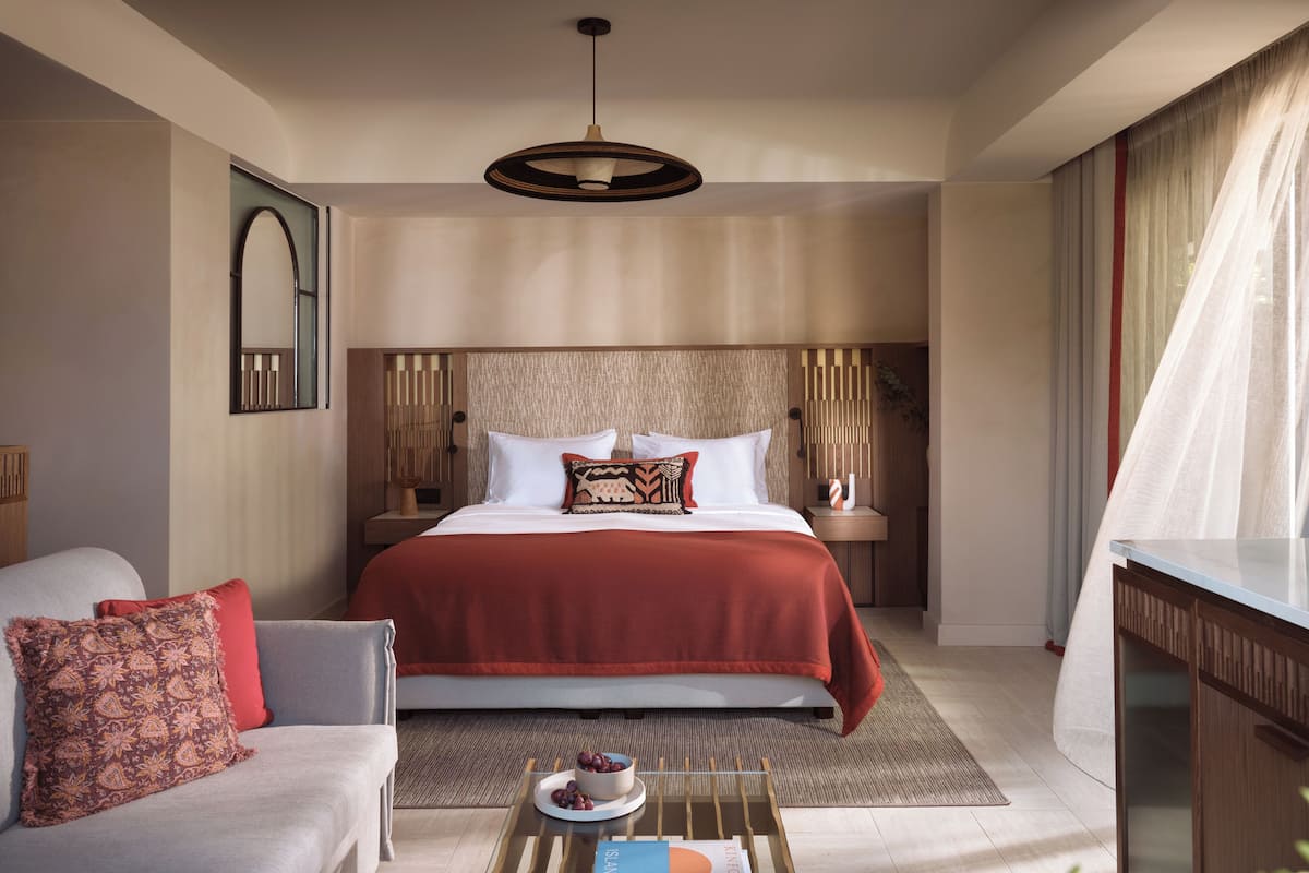 A modern hotel room with a large bed, a sofa, a bedside table with a lamp, and a view of the outdoors.