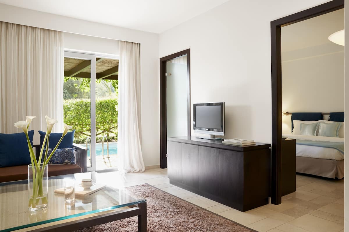 A modern hotel room with a glass dining table, a sofa, a TV on a dark wooden cabinet, and a large mirror.