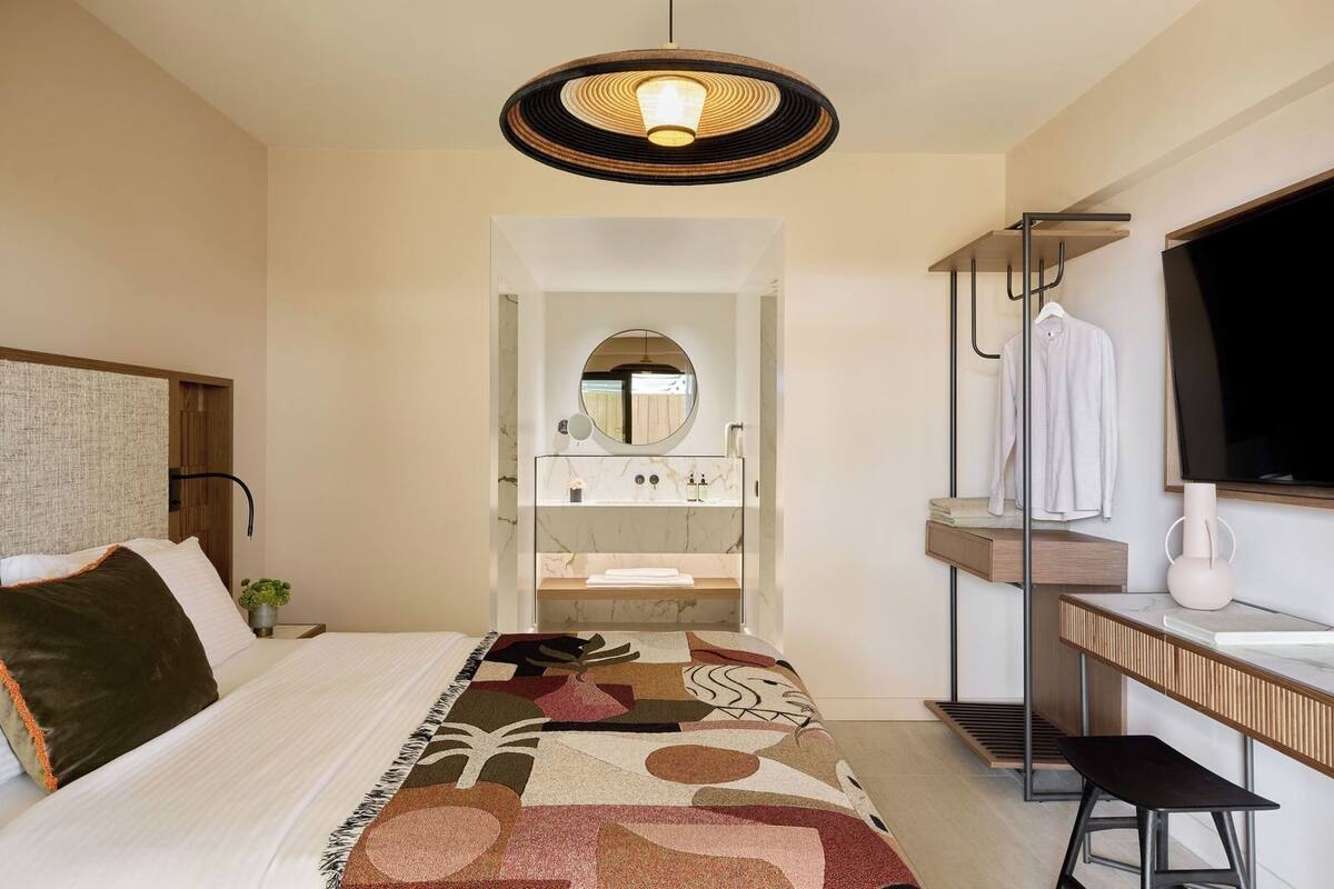 A modern hotel room with a bed, a large mirror, a vanity with a sink, and a television.